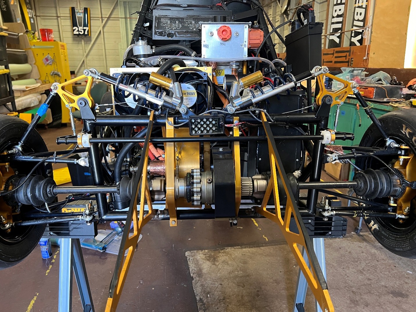Front view of an exposed automotive chassis on a lift, showing suspension arms, driveshafts, belts, and engine components in a workshop.